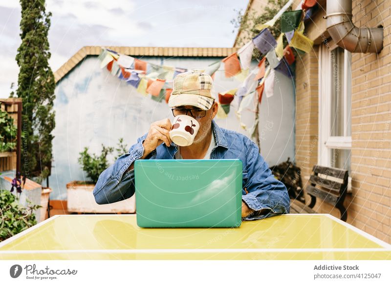 Nachdenklicher älterer Mann in Gläsern mit heißem Getränk und Laptop auf der Terrasse eines Landhauses trinken besinnlich lesen zuschauen verwenden Becher