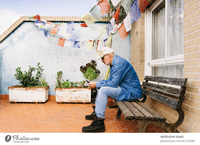 Nachdenklicher grauhaariger Mann in Jeanskleidung benutzt sein Smartphone, während er allein im Garten chillt Surfen Terrasse Kälte Nachricht lesen