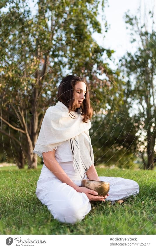 Konzentrierte junge Frau mit tibetischer Schale auf grüner Wiese Yoga tibetische Schale meditieren Klangschale Asana Windstille Zen Rasen Pose Sommer Knie Baum
