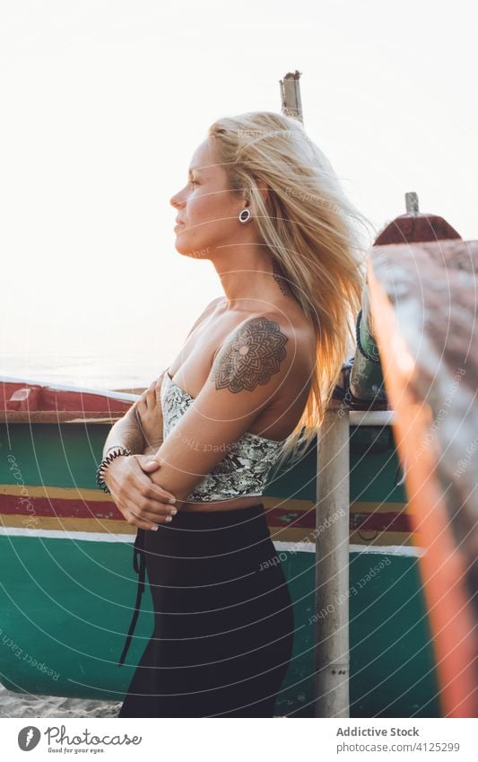 Tätowierte Frau steht mit verschränkten Armen in der Nähe eines Bootes am Sandstrand Strand Stil tropisch Sonnenuntergang Tattoo schlank Hand erhoben Kopf Ufer