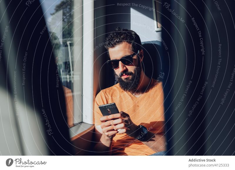 Ernster bärtiger Mann mit Sonnenbrille, der im Zug auf seinem Smartphone surft Passagier Arbeitsweg reisen Sommer Mobile Telefon Browsen Verkehr Fenster