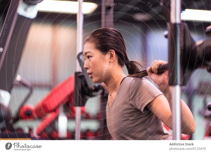 Kräftige junge asiatische Sportlerin macht Kniebeuge mit Langhantel in moderner Turnhalle Frau Curl-Hantel Training aktive Kleidung Maschine Gerät Fokus atmen