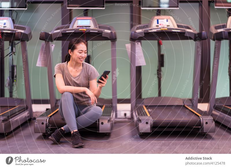 Fröhliche junge ethnische Sportlerin auf dem Laufband sitzend und ein Mobiltelefon im Fitnessstudio benutzend Frau Smartphone ruhen Pause Handy Training Herz