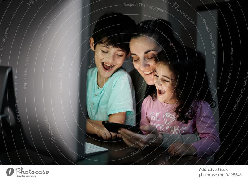 Fröhliche Frau mit Kindern schaut Video am Computer an Mutter benutzend Smartphone Zusammensein Telearbeit Kinderbetreuung Glück heiter heimwärts Kommunizieren