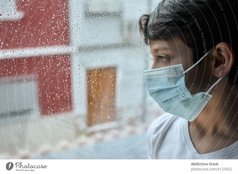 Trauriger Junge mit medizinischer Maske am Fenster Mundschutz Coronavirus Seuche heimwärts traurig unglücklich Selbstisolierung soziale Distanzierung Kind
