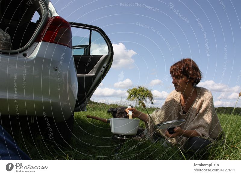 Frau sitzt im Grünen neben einem Auto und rührt in einem Kochtopf kochen Ausflug Ausflug ins Grüne Freiheit Trip Roadtrip reisen Picknick Natur genießen