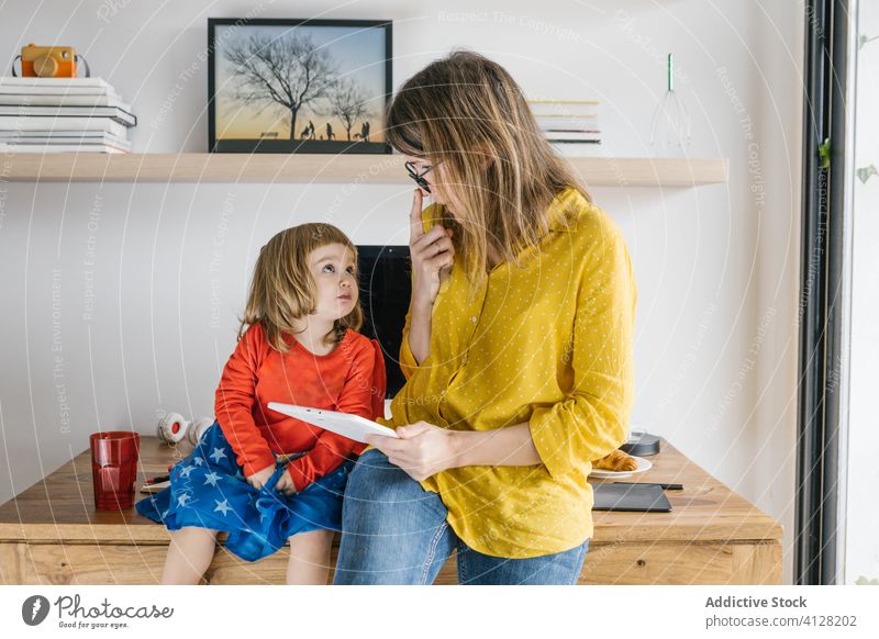 Glückliche Mutter und Tochter nehmen zu Hause Tabletten Frau freiberuflich Kind teilen Telearbeit Schreibtisch benutzend Apparatur Zusammensein Mädchen Brille