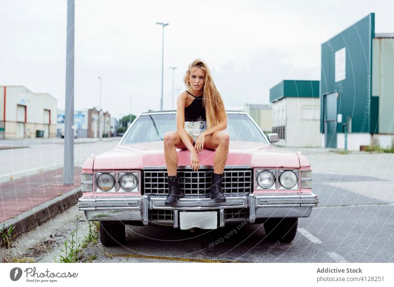 Attraktives blondes Mädchen sitzt auf der Motorhaube eines klassischen rosa Autos auf der Straße Frau jung Zopf Sitzen PKW alt Grunge Sommer Porträt Freizeit