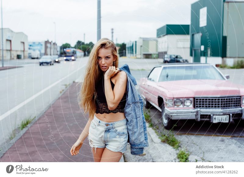 Sinnliches blondes Mädchen, das auf dem Bürgersteig steht und in die Kamera schaut, in der Nähe eines klassischen rosa Autos Frau jung Zopf Sitzen PKW alt