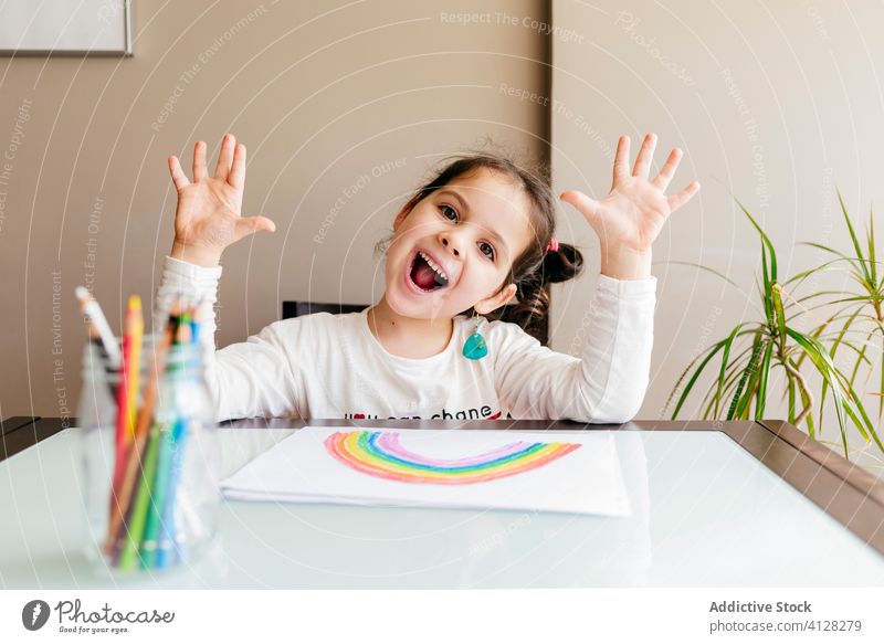 Fröhliches kleines Mädchen schaut in die Kamera, während es zu Hause zeichnet positiv Spaß Anstreicher Zeichnung hochreichen Handfläche spielerisch Grimasse