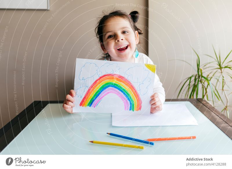 Fröhliches Mädchen zeigt der Kamera eine Zeichnung heiter farbenfroh zeigen Regenbogen positiv Optimist charmant Tisch zeichnen Papier Kind Lächeln niedlich