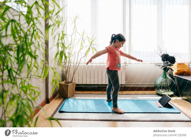 Fröhliches kleines Mädchen macht Yoga im hellen Raum Kind Pose Unterlage Video Tablette Tutorial Gleichgewicht üben zu Hause Stressabbau zuschauen Harmonie