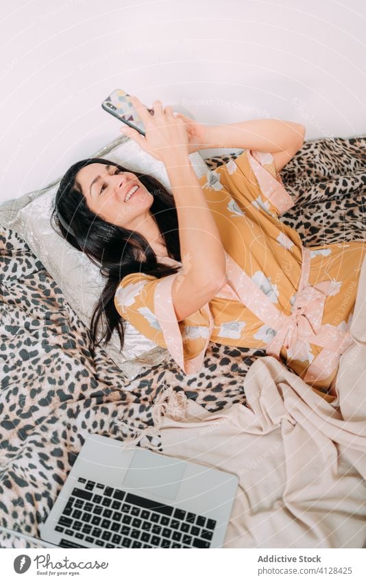 Lächelnde Frau surft auf dem Bett auf ihrem Smartphone Seide Robe Kälte soziale Netzwerke Komfort Kommunizieren benutzend sich[Akk] entspannen Internet Browsen
