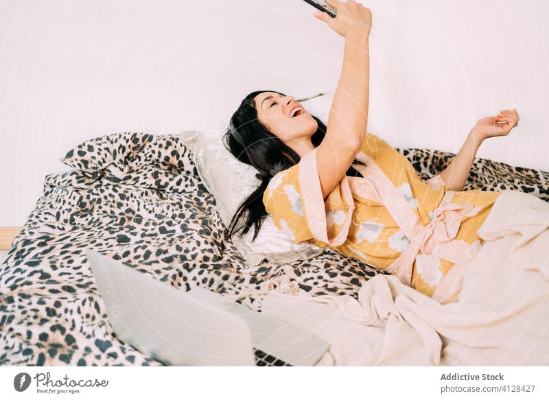 Frau singt auf dem Bett und hört Musik auf ihrem Smartphone Gesang Seide Robe Kälte Kopfhörer soziale Netzwerke Komfort Kommunizieren benutzend