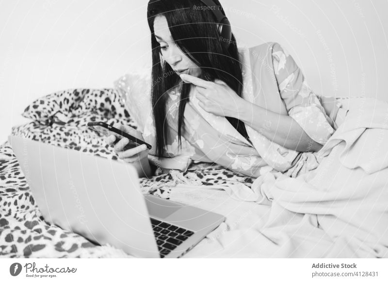Frau surft auf ihrem Smartphone, während sie im Bett Musik hört Seide Robe Kälte Kopfhörer soziale Netzwerke Komfort Kommunizieren benutzend