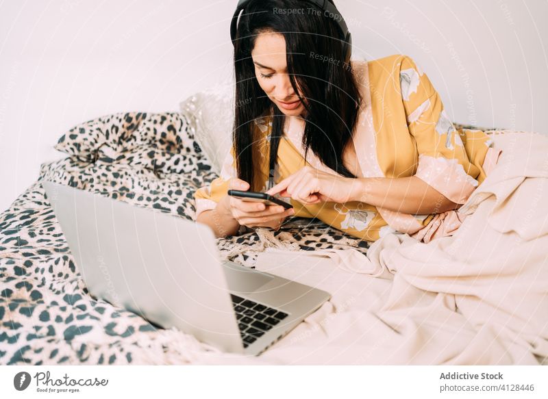 Frau surft auf ihrem Smartphone, während sie im Bett Musik hört Seide Robe Kälte Kopfhörer soziale Netzwerke Komfort Kommunizieren benutzend