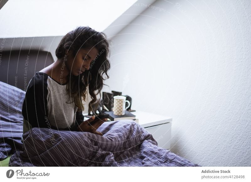 Junge ethnische Frau auf einem bequemen Bett, die ein Smartphone benutzt nachdenklich Inder Nachtwäsche Depression Problematik Komfort weich gemütlich
