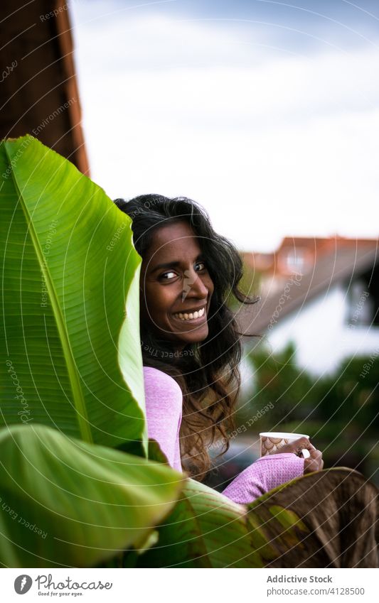 Lächelnde junge ethnische Frau mit einer Tasse Heißgetränk auf einer Terrasse positiv Balkon Kaffee Morgen ruhen warm Getränk trinken schön heiter