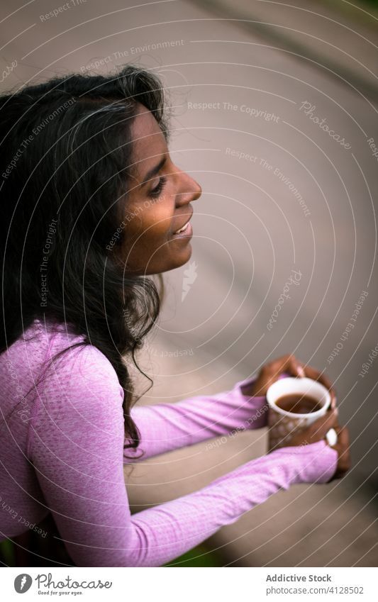 Lächelnde junge ethnische Dame mit einer Tasse Heißgetränk auf der Terrasse Frau positiv Balkon Kaffee Morgen ruhen warm Getränk rosa trinken schön heiter
