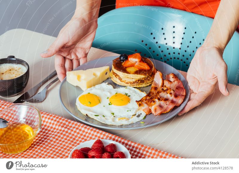 Anonymer Erntehelfer stellt Teller mit leckeren Spiegeleiern mit Speck und Käse auf den Frühstückstisch Person Hand Ei Muffins erdbeeren schwarze Beeren