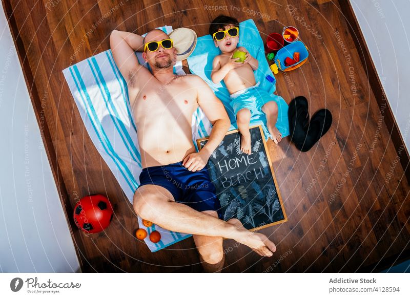 Vater und Sohn mit Sonnenbrille ruhen in Badehose auf dem Boden Strand heimwärts Selbstisolierung Quarantäne Sonnenbad träumen vorwegnehmen