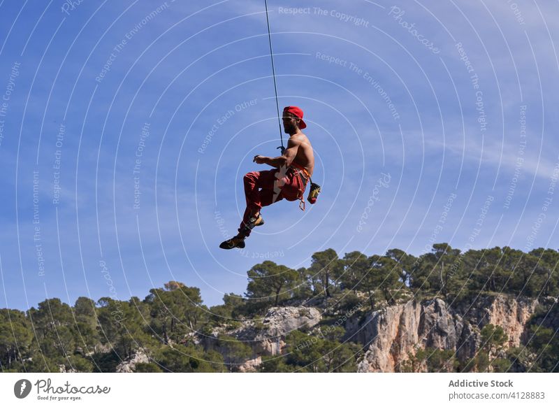 Starker männlicher Kletterer hängt an einem Seil in der Nähe eines felsigen Berghangs an einem sonnigen Tag Mann Bergsteiger stark Sport hängen sicheres Gerät