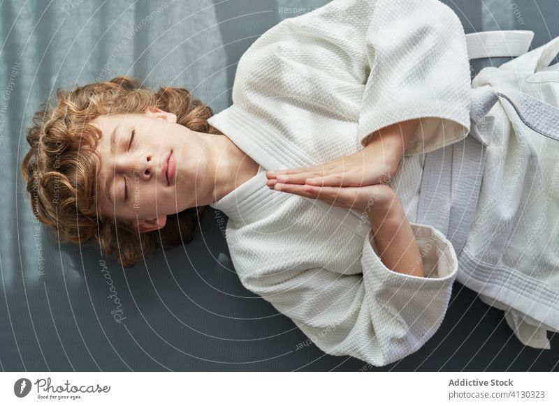 Konzentrierter Junge im weißen Kimono beim Meditieren üben liegend meditieren Judo Kind heimwärts Augen geschlossen Hände zusammen Übung Windstille