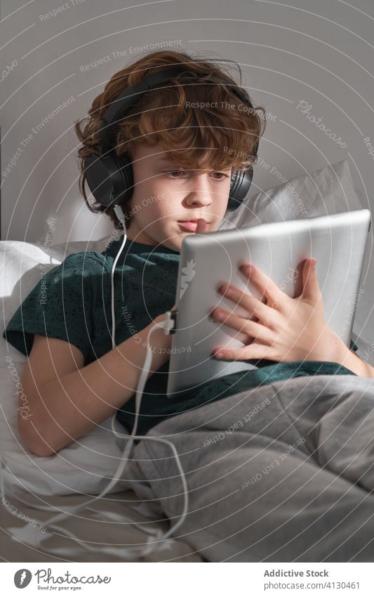 Zufriedenes Kind mit Kopfhörern, das ein digitales Tablet benutzt, während es in einem modernen Schlafzimmer zu Hause seinen freien Tag verbringt Tablette