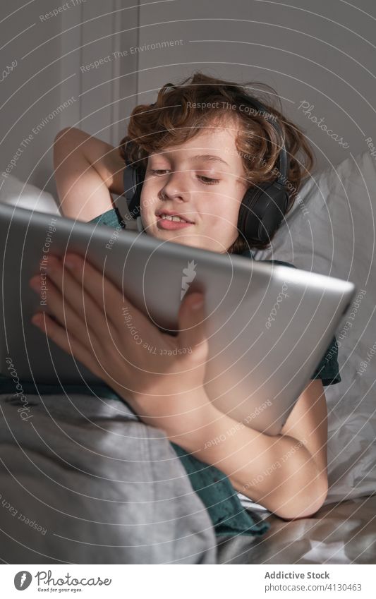 Zufriedenes Kind mit Kopfhörern, das ein digitales Tablet benutzt, während es in einem modernen Schlafzimmer zu Hause seinen freien Tag verbringt Tablette