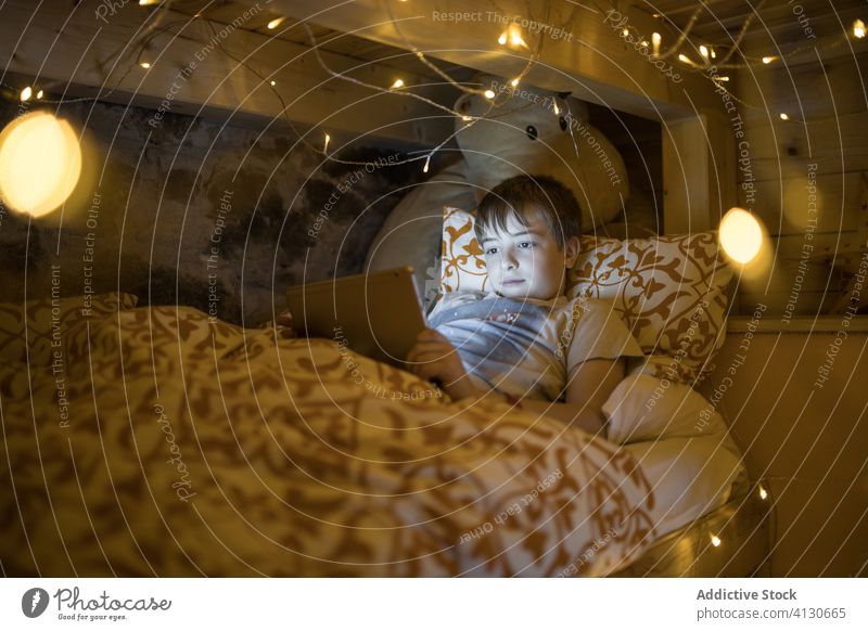 Kind schaut Tablette im Bett mit Girlande dekoriert Junge benutzend gemütlich Komfort zuschauen Video Kälte Freizeit heimwärts Abend glühen leuchten