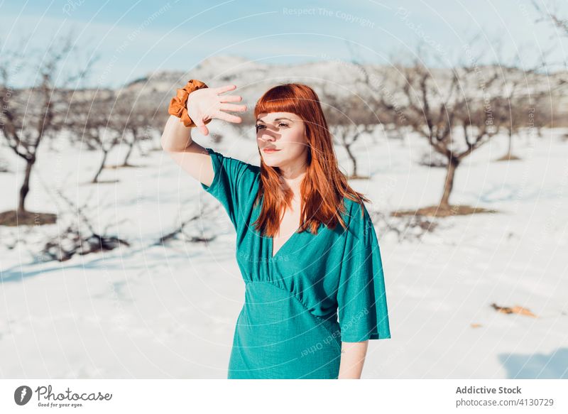 Junge schöne Frau mit langen roten Haaren in türkisem Outfit schaut weg, während sie in einem verschneiten Feld an einem sonnigen Tag steht Stil trendy sinnlich