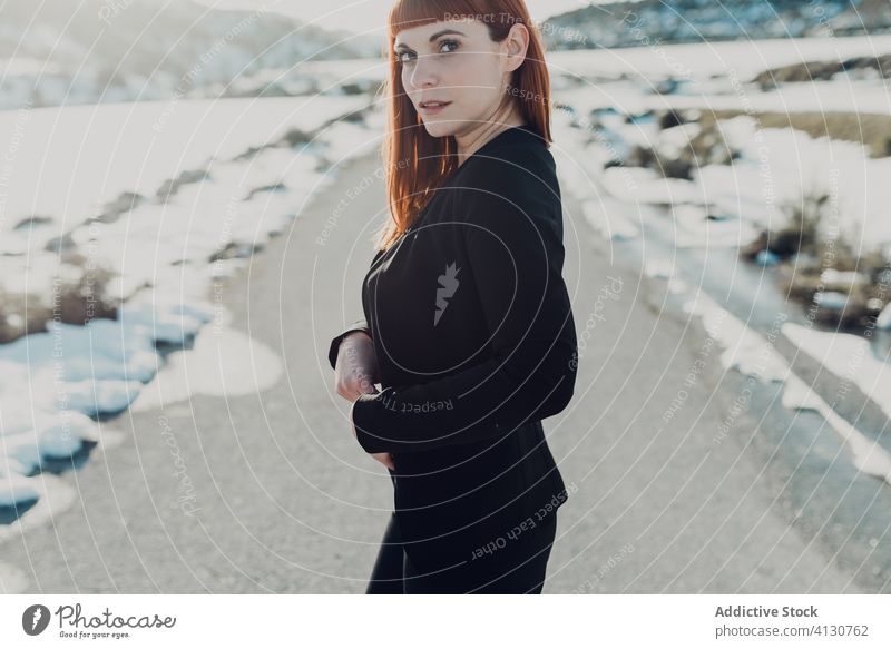 Junge Frau in schwarzer Kleidung steht auf verschneiter Straße Stil trendy Landschaft Anzug Natur Schnee Feld Berge u. Gebirge jung Winter reisen Lächeln Mode
