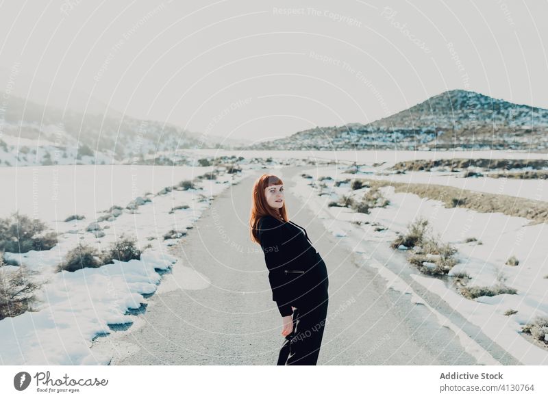 Junge Frau in schwarzer Kleidung steht auf verschneiter Straße Stil trendy Landschaft Anzug Natur Schnee Feld Berge u. Gebirge jung Winter reisen Lächeln Mode