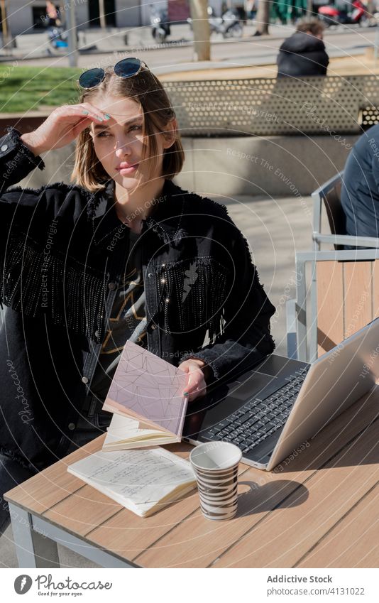 Studentin mit Laptop in einem Straßencafé im Sonnenlicht sitzend Freiberufler Schönschreibheft Augen abdecken hell Brille Kaffee Pause Freizeit Internet