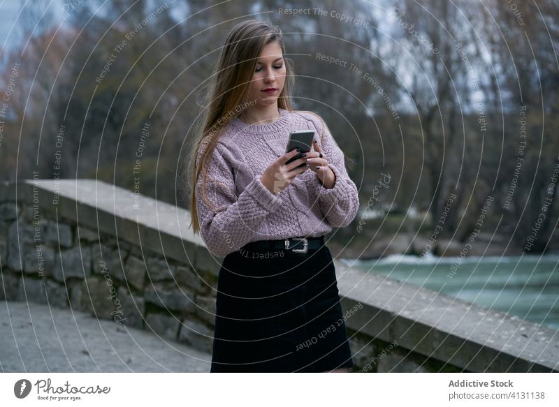 Konzentrierte junge Frau, die während eines Abendspaziergangs mit dem Telefon Nachrichten austauscht Nachrichtenübermittlung Park Herbst schlendern trendy