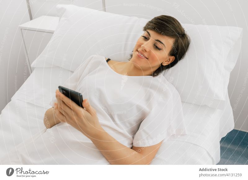 Glückliche Patientin mit Smartphone auf der Krankenhausstation Frau geduldig Station liegend benutzend Lächeln Stuhl Bett modern Robe heiter weiß Browsen Klinik