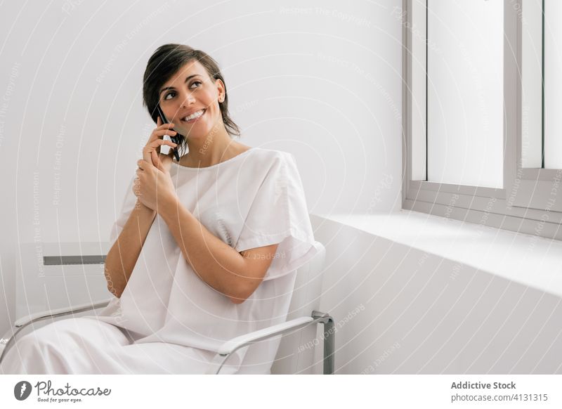 Glücklicher Patient, der in einer Krankenhausabteilung mit einem Smartphone spricht Frau geduldig Station sprechen benutzend Lächeln reden Stuhl sitzen Bett