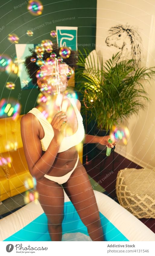 Schwarze Frau bläst Seifenblasen im Wohnzimmer Schlag Pool zu Hause bleiben soziale Distanzierung aufblasbar Schaumblase Selbstisolierung heimwärts Wochenende