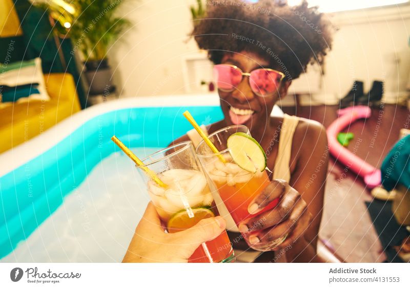 Fröhliche ethnische Frau stößt mit einem Freund auf ein Cocktailglas an Klirren Pool zu Hause bleiben Glas Alkohol trinken aufblasbar Selbstisolierung