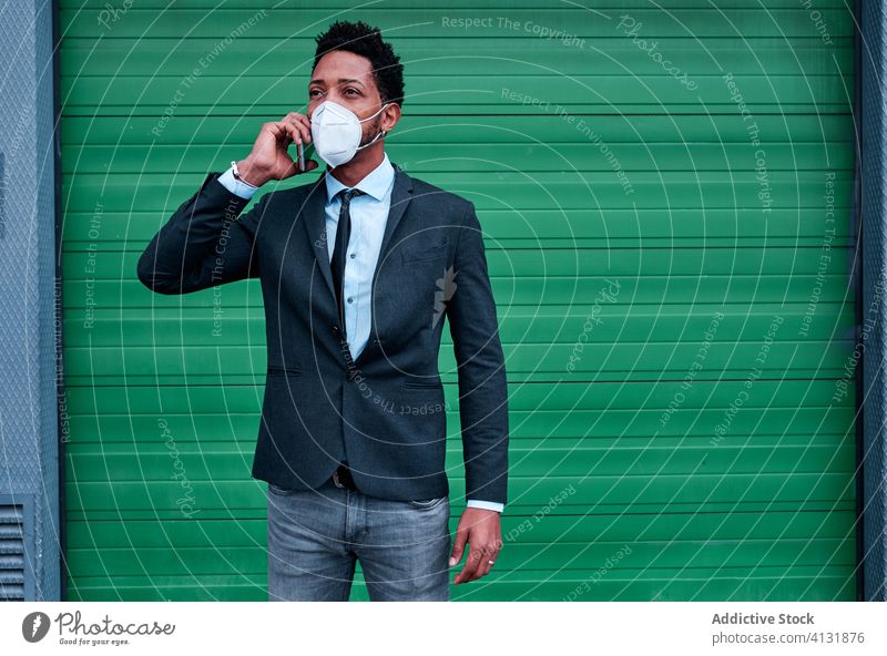 Seriöser ethnischer Geschäftsmann mit Atemschutzgerät spricht mit Smartphone auf der Straße Telefonanruf Mundschutz behüten Großstadt reden Pandemie Infektion