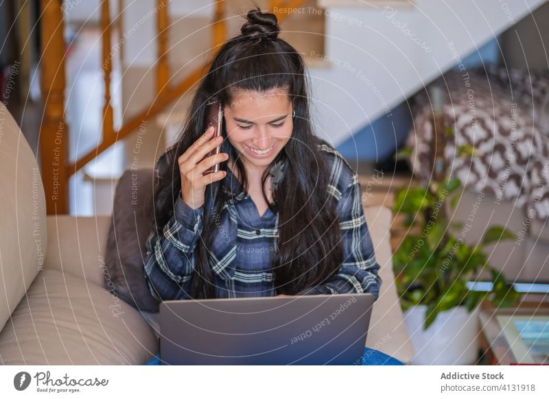 Positive Frau mit Laptop, die zu Hause mit ihrem Smartphone spricht heimwärts reden benutzend heiter jung Anruf Apparatur lässig freiberuflich Mobile Telefon