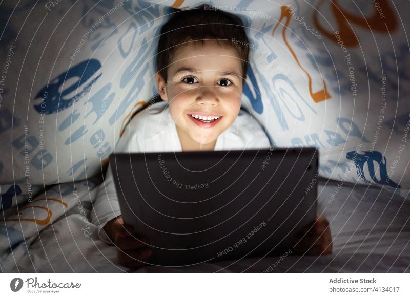 Entzückter Junge sieht Zeichentrickfilm auf dem Tablett Kind benutzend Tablette Decke Nacht zuschauend heiter Karikatur Pyjama heimwärts bezaubernd Gerät