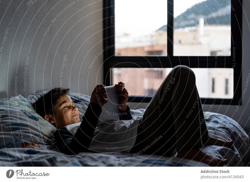 Entzückter Junge benutzt Tablette am Wochenende im Schlafzimmer Kind benutzend Decke Nacht zuschauend heiter Pyjama heimwärts bezaubernd Gerät Apparatur