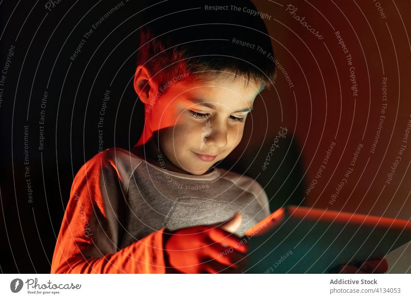 Entzückter Junge sieht Zeichentrickfilm auf dem Tablett Kind benutzend Tablette Nacht zuschauend heiter Karikatur Pyjama heimwärts bezaubernd Gerät Apparatur