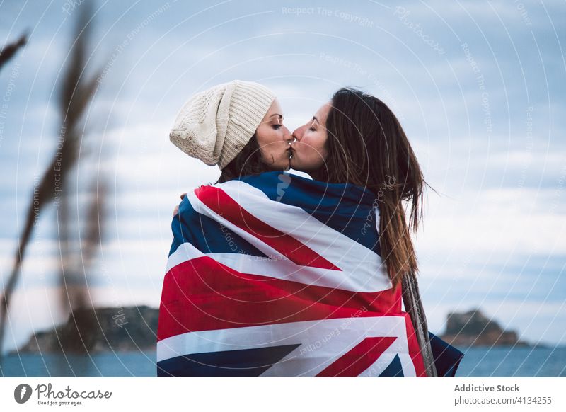Küssendes junges lesbisches Paar mit britischer Flagge Großbritannien Fahne Meeresufer wolkig Himmel Kuss Umarmung umhüllen Frauen Augen geschlossen Hut