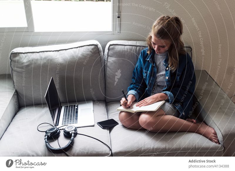 Millennial-Frau sitzt auf dem Sofa mit Gadgets und schreibt in ein Notizbuch schreiben Notebook heimwärts Apparatur jung zur Kenntnis nehmen Tagebuch