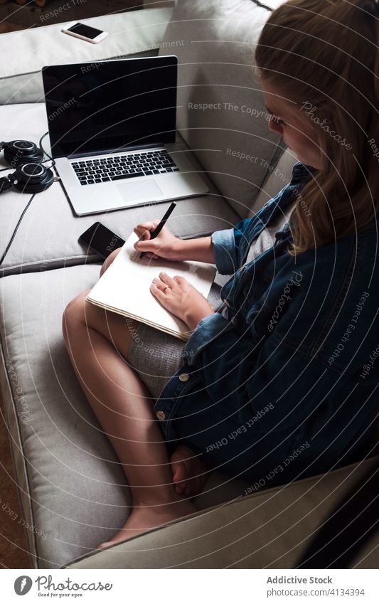 Millennial-Frau sitzt auf dem Sofa mit Gadgets und schreibt in ein Notizbuch schreiben Notebook heimwärts Apparatur jung zur Kenntnis nehmen Tagebuch
