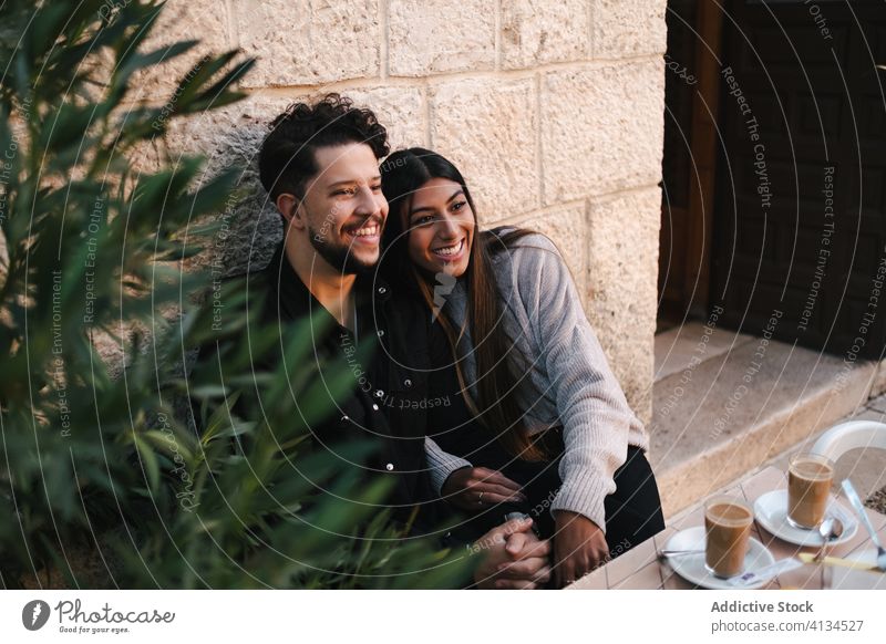 Zufriedenes multiethnisches Paar auf der Straße sitzend und lachend Lachen Umarmung Zusammensein heiter schlendern Großstadt Spaß haben Liebe rassenübergreifend