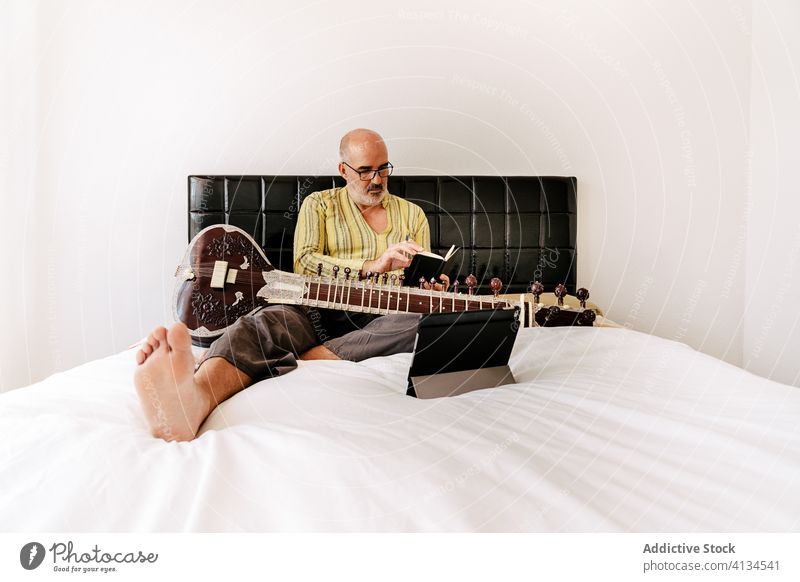 Älterer männlicher Musiker, der zu Hause Sitar spielt sitar Mann spielen Tablette benutzend online Lektion älter Notebook zur Kenntnis nehmen Apparatur Gerät