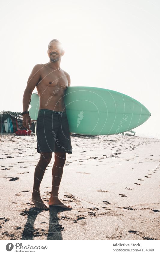 Fröhlicher Surfer mit Surfbrett am Strand stehend Mann Glück Strandpromenade aktiv Meer Küste Lächeln ohne Hemd ethnisch männlich Lifestyle Sand Sommer Ufer